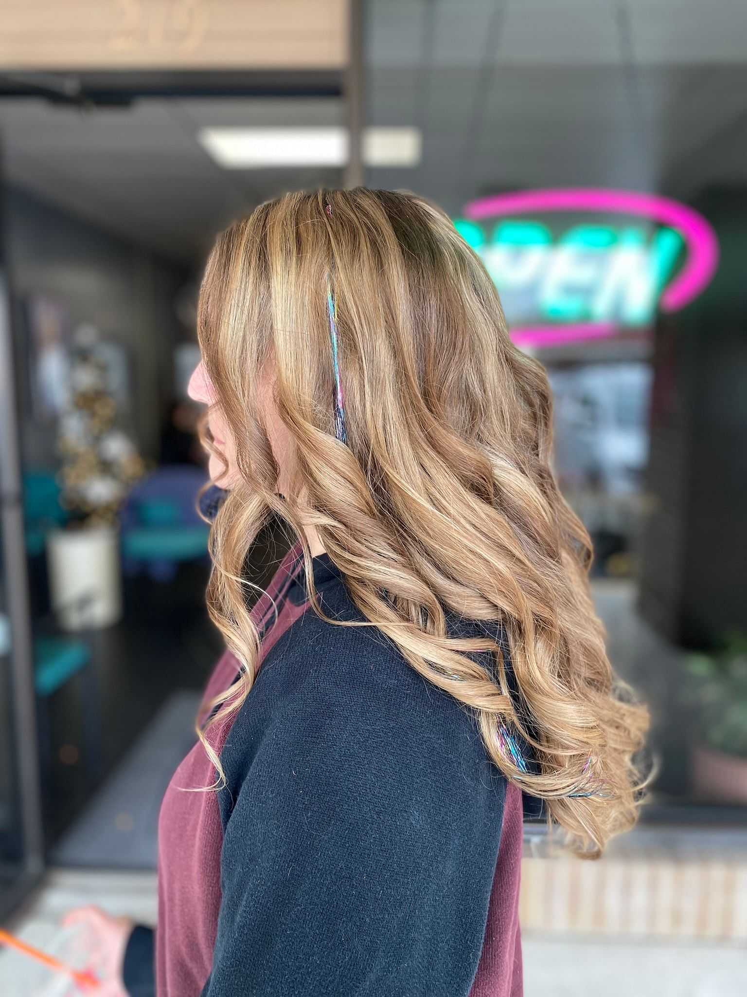 Woman with wavy blonde hair and colorful hair streaks standing outside a salon with a neon "OPEN" sign.