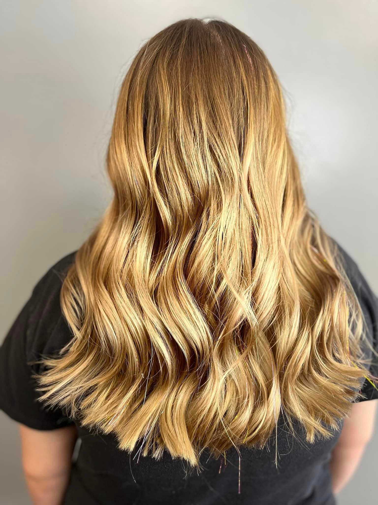 Back view of a person with long, wavy blonde hair wearing a black shirt against a plain background.